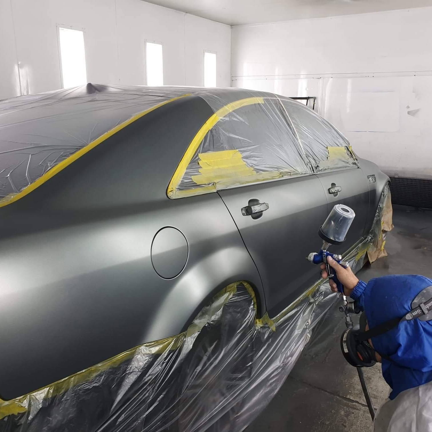A Pro Panel and Mechanical painter spraying a masked-up grey sedan in the spray booth
